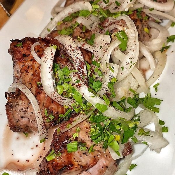 Grilled Georgian shashlik, topped with sliced onions, fresh herbs, and red spice on a white plate.